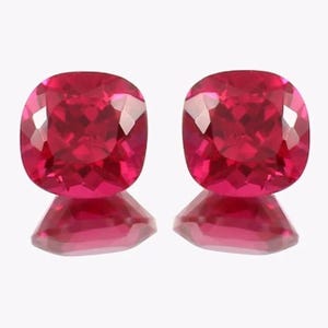 Könnte beinhalten: Zwei kissenförmig geschliffene, facettierte, rote Edelsteine. Die Edelsteine haben eine leuchtende, tiefrote Farbe und werden auf einer reflektierenden Oberfläche präsentiert. Die Facetten fangen das Licht ein und erzeugen einen funkelnden Effekt.