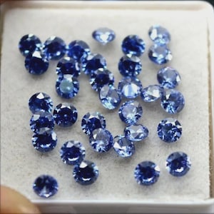 Op de afbeelding: Een verzameling ronde, gefacetteerde blauwe edelstenen, waarschijnlijk saffieren, gepresenteerd in een witte tray. De edelstenen variëren enigszins in grootte en tint, met een heldere, reflecterende kwaliteit.