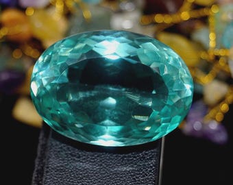May include: A large, faceted, oval-shaped gemstone in shades of teal and turquoise. The gem is set against a dark background, with a blurred backdrop of other colorful stones and gold accents.