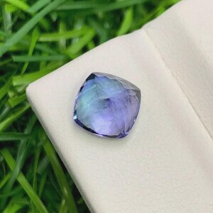 Pu&ograve; includere: Una gemma taglio cuscino con sfumature blu e viola. La gemma &egrave; posta su uno sfondo bianco, con erba verde visibile sullo sfondo. Le sfaccettature della pietra riflettono la luce, creando un effetto scintillante.