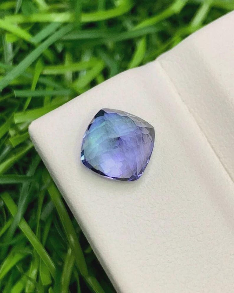 Pu&ograve; includere: Una gemma taglio cuscino con sfumature blu e viola. La gemma &egrave; posta su uno sfondo bianco, con erba verde visibile sullo sfondo. Le sfaccettature della pietra riflettono la luce, creando un effetto scintillante.