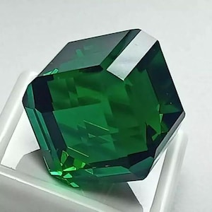 May include: A faceted, emerald-green gemstone with an octagonal shape. The gem is highly polished, reflecting light and creating a brilliant display of colour. The gemstone is set on a white surface.