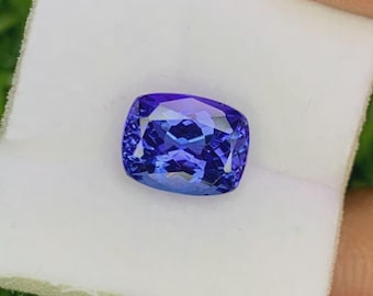 May include: A square-cut, blue gemstone with multiple facets. The gem is set on a white surface, with a green background. The facets of the gemstone reflect light, creating a sparkling effect. The gem is a vibrant, eye-catching piece.
