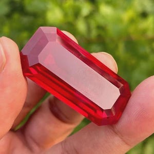 Könnte beinhalten: Ein facettierter, rechteckiger roter Edelstein, der in einer Hand gehalten wird. Der Edelstein hat eine polierte, reflektierende Oberfläche und eine tiefrote Farbe. Die Form des Edelsteins ist ein langes Rechteck mit abgewinkelten Kanten.