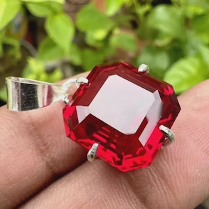 以下が含まれることがあります： 銀色のベイルが付いた、ファセット加工された八角形の深紅色の宝石ペンダント。 宝石は4つの小さな銀色の爪で固定されています。 ぼやけた緑色の背景にペンダントが表示されています。