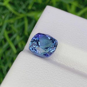 Pu&ograve; includere: Una gemma taglio cuscino con una sfumatura di colore blu e verde acqua. La gemma &egrave; posta su una superficie bianca, con uno sfondo verde sfocato. Le sfaccettature della pietra riflettono la luce, creando un effetto scintillante. Questa gemma &egrave; probabilmente per gioielli.