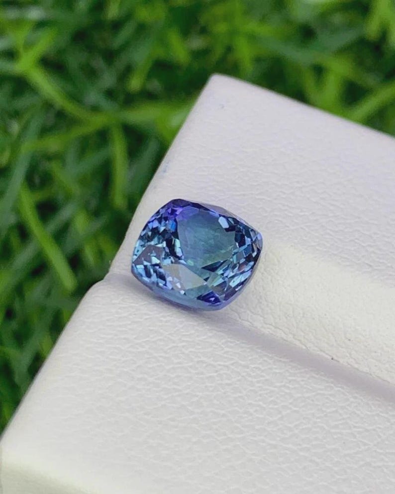 Pu&ograve; includere: Una gemma taglio cuscino con una sfumatura di colore blu e verde acqua. La gemma &egrave; posta su una superficie bianca, con uno sfondo verde sfocato. Le sfaccettature della pietra riflettono la luce, creando un effetto scintillante. Questa gemma &egrave; probabilmente per gioielli.