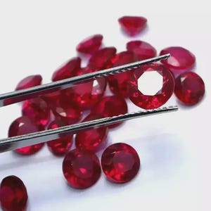 May include: A collection of faceted, round, deep red gemstones, resembling rubies, are scattered on a white surface. One gem is held by silver tweezers, showcasing its intricate cut and clarity. The gems vary slightly in size and shape, reflecting light.