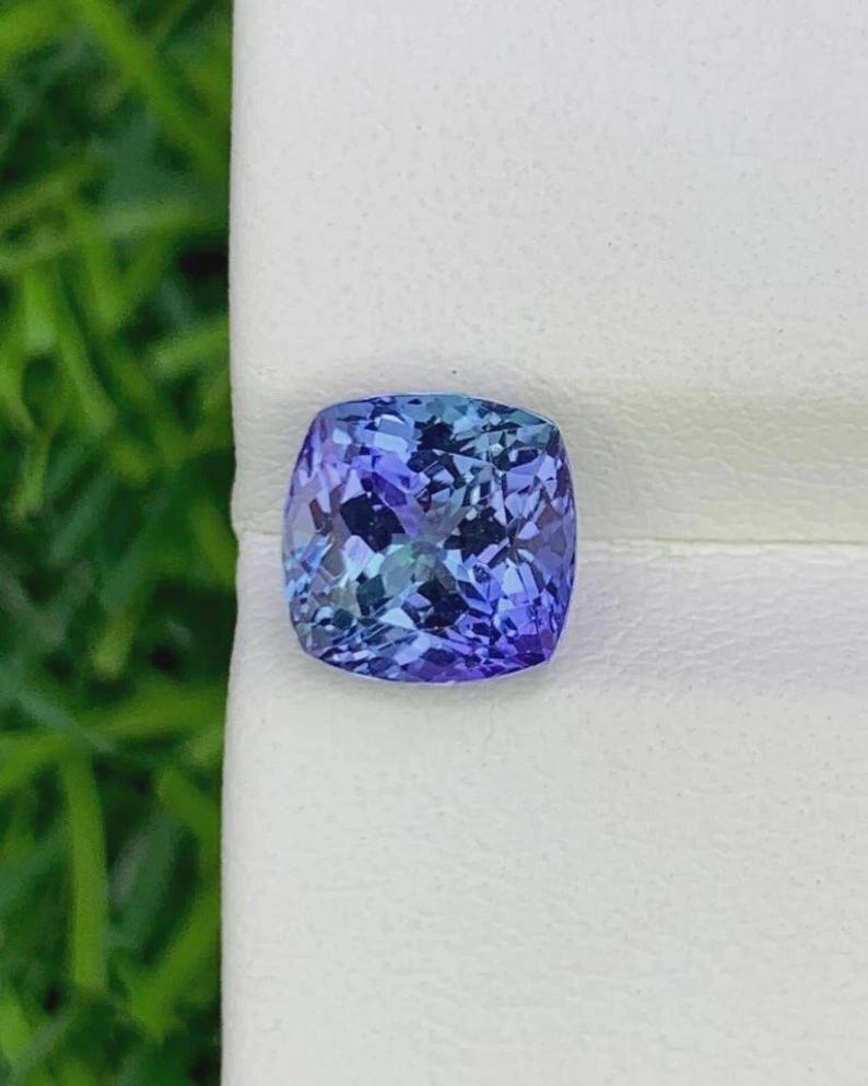 Pu&ograve; includere: Una gemma taglio cuscino con una vibrante gamma di blu e viola. Le sfaccettature della gemma riflettono la luce, creando un effetto scintillante. La pietra &egrave; posta su uno sfondo bianco, evidenziando il suo colore e la sua chiarezza. Un bellissimo esempio di alta gioielleria.