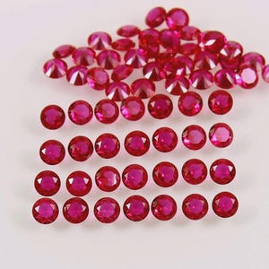 May include: A collection of round, faceted, deep red gemstones. The gems are arranged in rows and scattered across a white surface, showcasing their brilliant cut and colour. These appear to be rubies.