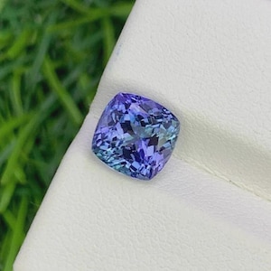 Pu&ograve; includere: Una gemma di tanzanite taglio a cuscino, che mostra una miscela di tonalit&agrave; blu e viola. La gemma &egrave; posta su una superficie bianca, con uno sfondo verde sfocato. Le sfaccettature della gemma riflettono la luce.