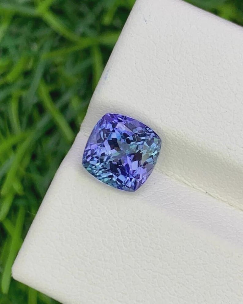 Pu&ograve; includere: Una gemma di tanzanite taglio a cuscino, che mostra una miscela di tonalit&agrave; blu e viola. La gemma &egrave; posta su una superficie bianca, con uno sfondo verde sfocato. Le sfaccettature della gemma riflettono la luce.