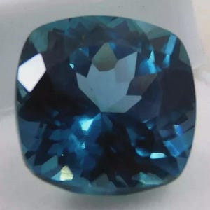 May include: A faceted, cushion-cut gemstone with a deep teal color. The gem's surface reflects light, creating a sparkling effect. The stone's shape is square with rounded corners, and it appears to be set against a white background.