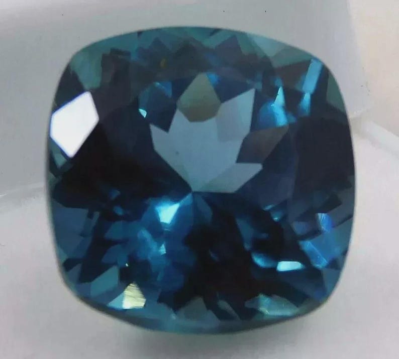 May include: A faceted, cushion-cut gemstone with a deep teal color. The gem's surface reflects light, creating a sparkling effect. The stone's shape is square with rounded corners, and it appears to be set against a white background.