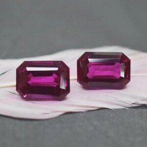 May include: Two rectangular, faceted gemstones with a deep magenta hue. The gems are displayed on a soft, pinkish-white surface against a gray backdrop. The cut of the stones allows light to reflect, creating a brilliant sparkle.