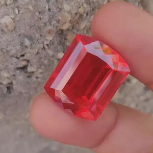 May include: A faceted, rectangular red gemstone with a brilliant cut. The gem is held between fingers, showcasing its vibrant colour and reflective surfaces. The gem's facets catch the light, creating a dazzling display.