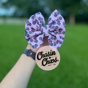 May include: A large, light purple hair bow with a repeating pattern of small motorcycles in black and purple. The bow is held in front of a blurred green background. A round sticker with the text "Chasin Chaos Co." is visible.
