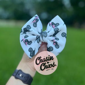 Op de afbeelding: Witte haarstrik met een patroon van zwarte motorfietsen en roze accenten. De strik wordt voor een groene achtergrond gehouden. Een roze cirkel met de tekst "Chasin Chaos Co." is zichtbaar.