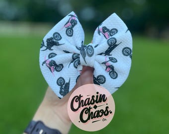 May include: A white fabric hair bow with a textured surface featuring a repeating pattern of black and pink dirt bikes. A circular pink tag is attached to the bow, displaying the black text Chasin Chaos Co. in a stylized script font.