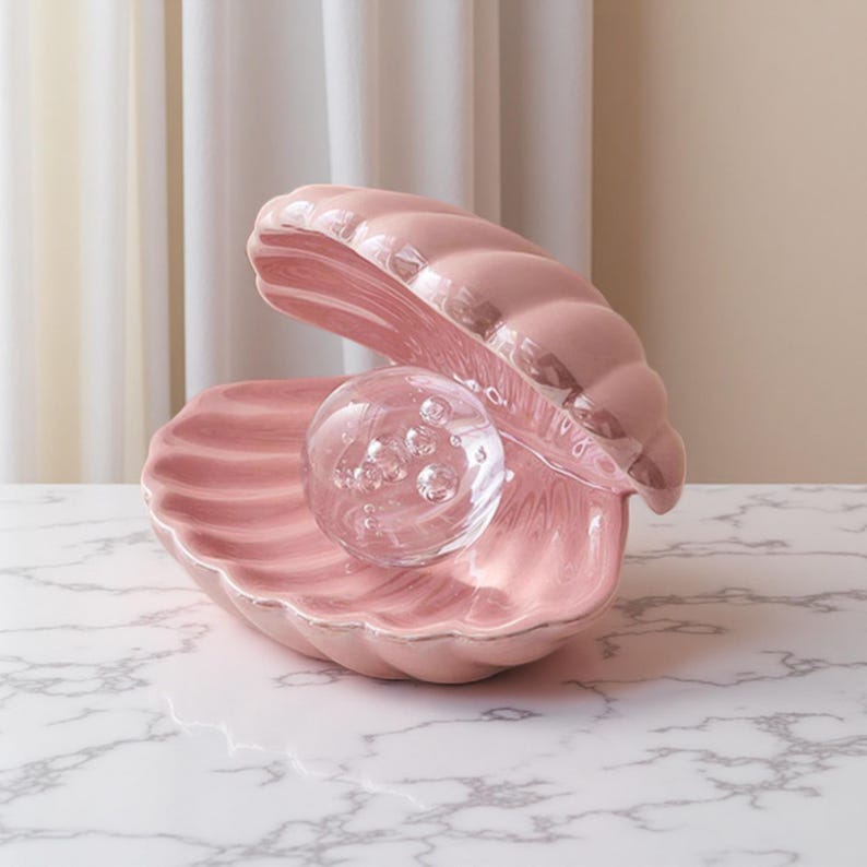 May include: A pink ceramic seashell-shaped dish with a clear glass sphere containing bubbles. The dish has a glossy finish. The background is a neutral colour. The dish is a decorative item.