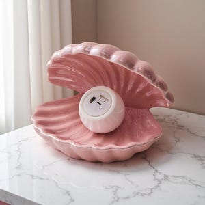 May include: A pink ceramic seashell-shaped lamp with a white, round light fixture inside. The lamp has a glossy finish and sits on a marble-patterned surface. The lamp is a decorative lighting fixture.