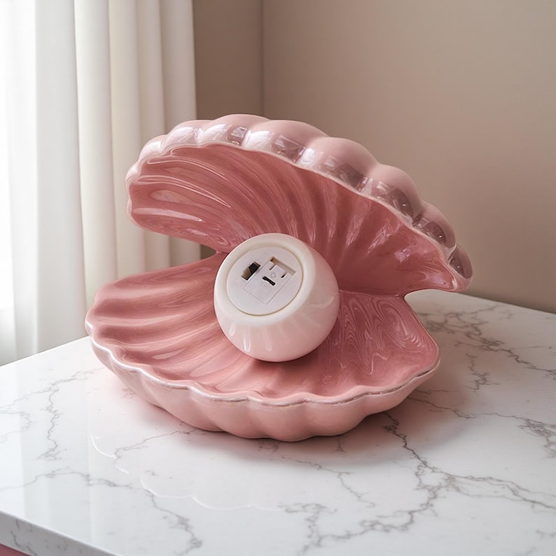 May include: A pink ceramic seashell-shaped lamp with a white, round light fixture inside. The lamp has a glossy finish and sits on a marble-patterned surface. The lamp is a decorative lighting fixture.