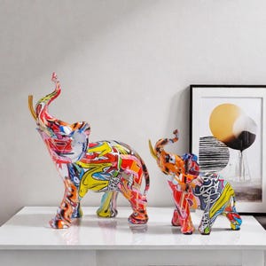 Peut inclure: Deux sculptures d'éléphants colorés avec des motifs de style graffiti. Le plus grand éléphant mesure environ 30 cm de haut, le plus petit environ 20 cm. Les sculptures présentent un mélange vibrant de rouge, jaune, bleu et blanc.