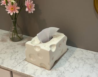 May include: A cream-coloured ceramic tissue box designed to resemble a block of Swiss cheese, complete with holes. A tissue is visible. The box is on a marble-effect surface, with pink flowers in a vase behind it.