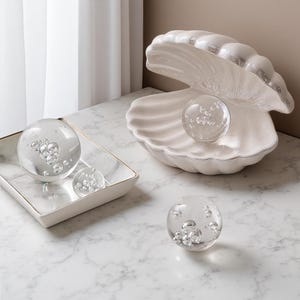 May include: A decorative arrangement featuring a large white ceramic seashell with a pearl-like glass sphere inside. Additional glass spheres with bubble inclusions are displayed on a marble tray and surface, creating a coastal theme.
