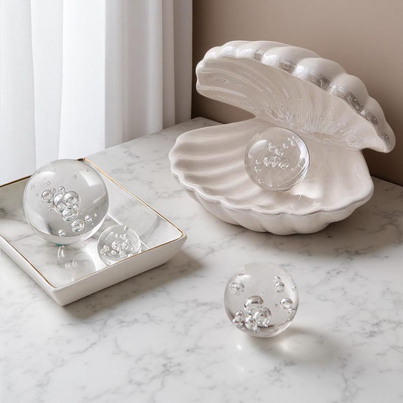 May include: A decorative arrangement featuring a large white ceramic seashell with a pearl-like glass sphere inside. Additional glass spheres with bubble inclusions are displayed on a marble tray and surface, creating a coastal theme.
