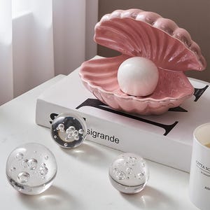May include: A pink ceramic seashell-shaped dish with a white pearl inside. Three clear glass spheres with air bubbles are in front of the dish. The dish sits on a white book with the word "sigrande" printed on it.
