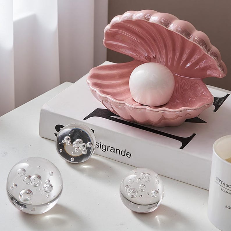 May include: A pink ceramic seashell-shaped dish with a white pearl inside. Three clear glass spheres with air bubbles are in front of the dish. The dish sits on a white book with the word "sigrande" printed on it.