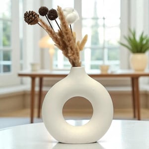 May include: A white ceramic vase with a circular opening, holding dried floral stems. The arrangement includes a pinecone, brown spherical buds, and fluffy white cotton-like elements. The vase sits on a white surface.