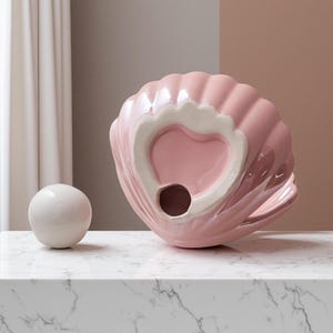 May include: A pink ceramic seashell-shaped decorative object with a white interior and a small hole. A white sphere sits to the left of the shell on a marble surface. The shell has a glossy finish.
