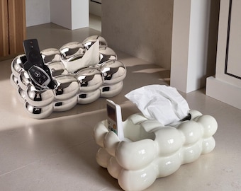 May include: Two decorative tissue box holders. One is silver and holds a remote and car keys. The other is white and holds a remote. Both have a bubble-like design and are filled with white tissues.