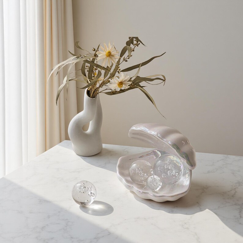 May include: A white ceramic vase with a distinctive, curved design holds dried flowers and greenery. A pearlescent, open clam shell dish contains three clear glass spheres. A single glass sphere sits nearby on a marble surface. Decorative home decor.