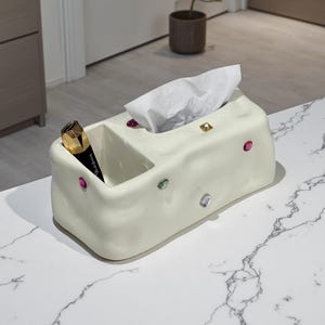 May include: A cream ceramic tissue box with a compartment for storage. The box is decorated with colourful, gem-like embellishments. A black and gold cosmetic tube sits in the compartment, and white tissues are in the opening.