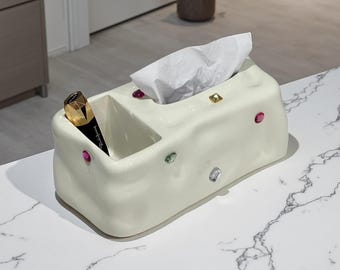 May include: A cream ceramic tissue box with a compartment for storage. The box is decorated with colourful, gem-like embellishments. A black and gold cosmetic tube sits in the compartment, and white tissues are in the opening.