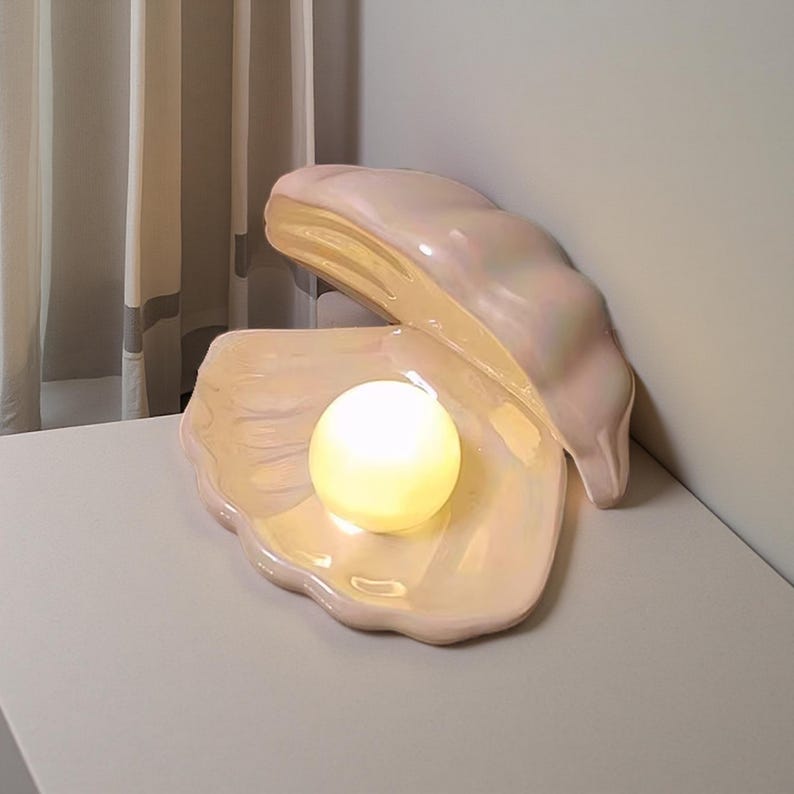 May include: A pearlescent pink clam shell lamp with a glowing spherical light inside. The lamp is open, revealing the light source, and sits on a neutral surface. The lamp's design is smooth and glossy, creating a soft, ambient glow.