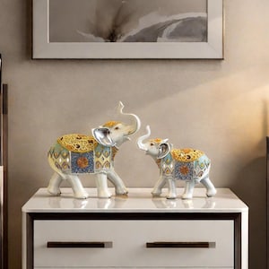 Puede incluir: Dos figuras decorativas de elefantes blancos con colmillos y trompas plateadas. Tienen mantas de silla ornamentadas y coloridas con diseños dorados, azules y naranjas. Las figuras se exhiben sobre una superficie blanca.