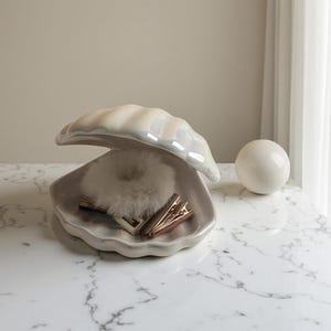 May include: A pearlescent ceramic clam shell-shaped trinket dish with a fluffy white scrunchie and two gold hair clips inside. A white ceramic sphere sits to the right. The dish is open, revealing its contents.