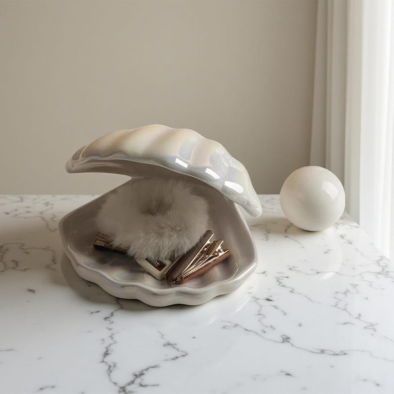 May include: A pearlescent ceramic clam shell-shaped trinket dish with a fluffy white scrunchie and two gold hair clips inside. A white ceramic sphere sits to the right. The dish is open, revealing its contents.
