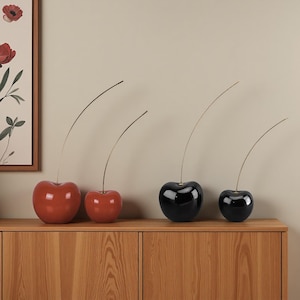 Peut inclure: Quatre sculptures décoratives en forme de cerises. Deux sont rouges, deux sont noires, toutes brillantes. Chaque cerise a une fine tige incurvée. Les cerises sont exposées sur un meuble en bois. Un cadre floral est visible en arrière-plan.