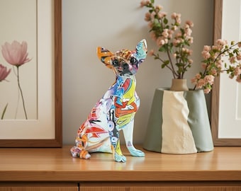 May include: A colorful Chihuahua figurine with a graffiti-style design. The dog statue features a mix of bright colors, including red, yellow, blue, and orange. The figurine is sitting on a wooden surface, with a vase and framed artwork in the background.