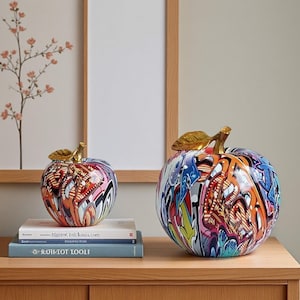 May include: Two decorative apple ornaments with graffiti-style patterns. The apples are multi-coloured with gold-coloured stems and leaves. One apple is smaller and sits on books. The other apple is larger and stands alone on a wooden surface.