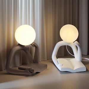 Puede incluir: Dos lámparas de mesa modernas, cada una con la forma de una persona leyendo. Una lámpara es de color topo, la otra blanca. Ambas tienen una bombilla esférica para la cabeza y la base es un libro. Las lámparas están sobre una superficie clara.