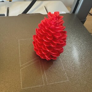 May include: A vibrant red, 3D-printed pinecone sits on a textured surface. The pinecone's detailed scales are clearly visible, showcasing the precision of the 3D printing process. The object is a decorative item, possibly for seasonal decor.