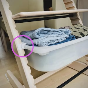 Può includere: Un seggiolone in legno chiaro con un vassoio in plastica bianca. Il vassoio contiene asciugamani blu e grigi piegati e un panno fantasia. Un pezzo di plastica bianca collega il vassoio alla sedia.