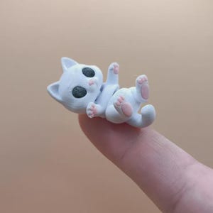 Könnte beinhalten: Eine kleine, weiße Katzenfigur mit schwarzen Augen und rosa Pfoten, die auf dem Rücken liegt und die Pfoten in die Luft reckt. Die Katze liegt auf einem Finger vor einem neutralen Hintergrund.