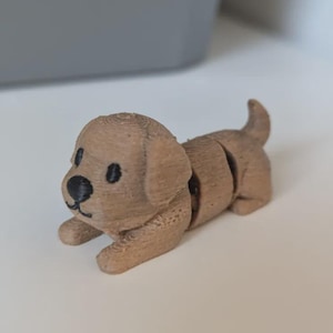 May include: A small, light brown 3D-printed dog figurine with black eyes and nose. The dog is segmented, allowing for movement. It is lying down on a white surface, with a gray object in the background.